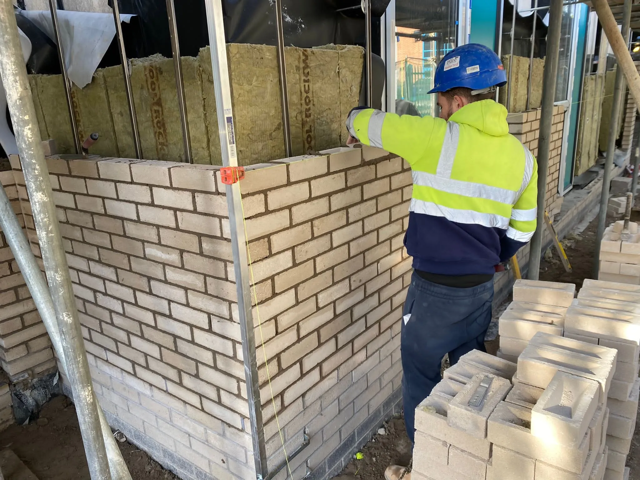 Construction worker wearing a blue hard hat and a high-visibility yellow and navy jacket laying bricks to build a corner wall on a construction site with insulation visible behind the metal framing.